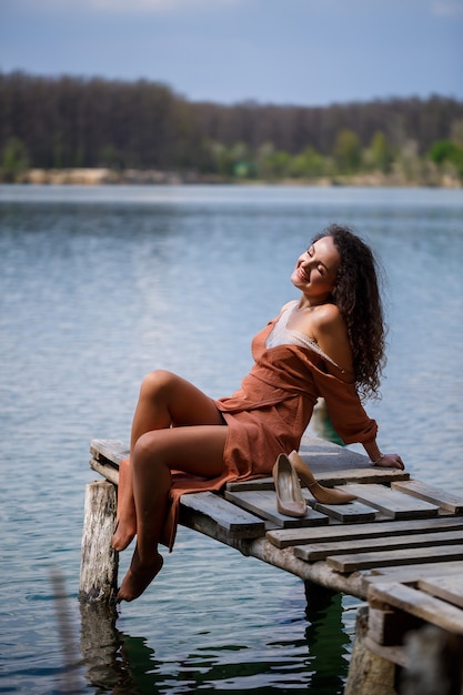 A girl with long wavy curly hair in a long guipure dress barefoot in the summer in a forest on a lake at sunset standing on a pantone on a wooden pier bridge. Summer sunny day