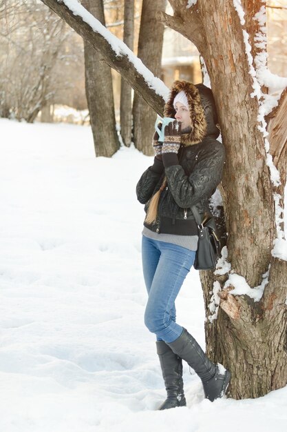 Girl with a mug of tea leaning against a tree and looking straight Winter outdoors