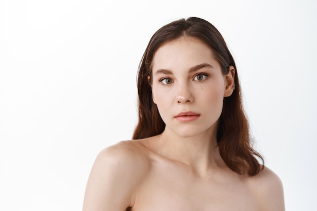 Girl with naked shoulders, hydrated clean facial skin, standing on white wall