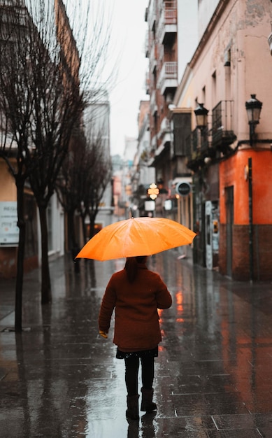 Girl with an orange umbrella protecting herself from the rain walking down a lonely narrow street