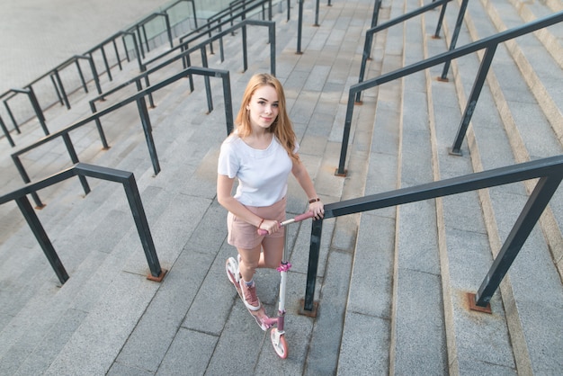 Girl with a pink kick scooter stands on the background of a modern architecture, view of the camera, top view.