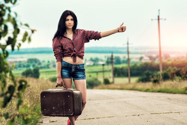 Girl with retro suitcase standing on hitchhiking. She dressed in shorts and a shirt stops the car.