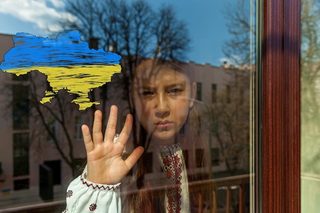 A girl with a sad face in an embroidered dress stands near the window with a drawing of a map