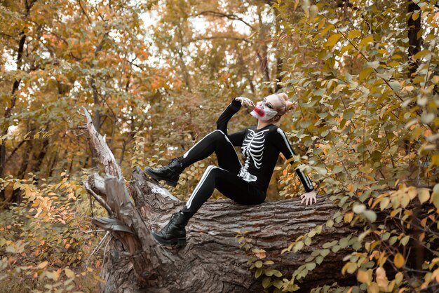 A girl with scary make-up in a skeleton costume sits on a withered old tree. Halloween