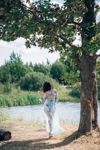 Girl with a sexy ass in a swimsuit and tunic poses on beach by river