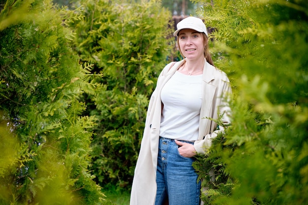 A girl with a smile on her face in jeans raincoat cap stands among the green bushes