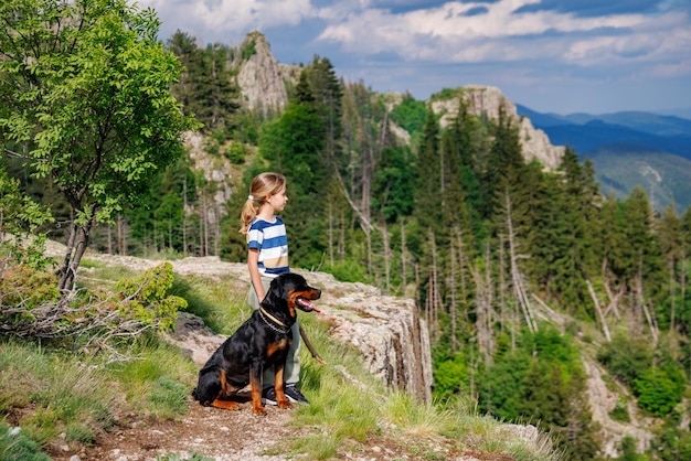 Girl with stands next to her dog of the Rottweiler breed on a peak with vegetation against the cloudy sky