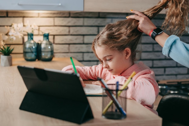 Girl with tablet pc computer writing to notebook while her mother arranges her hair distant educatio