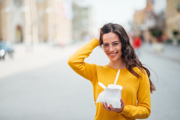 Girl with takeaway box standing in the city street, smiling at camera, copy space.