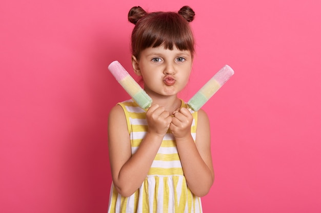 Girl with two water ice cream looking at camera and making grimace, pout lips, wearing striped summer dress, standing isolated over pink background.