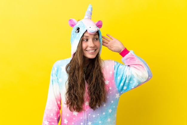 Girl with unicorn pajamas over isolated background listening to something by putting hand on the ear