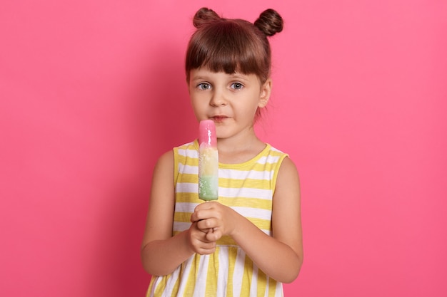 Girl with water ice cream looking directly at camera, posing isolated over rosy background, dresses striped white and yellow outfit, standing with funny knots, looks at camera.