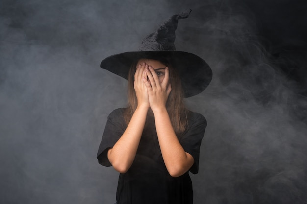 Girl with witch costume for halloween parties over isolated dark wall covering eyes and looking through fingers