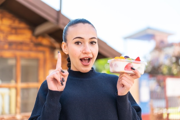 A girl with a yogurt bowl in her hand is holding up her index finger.