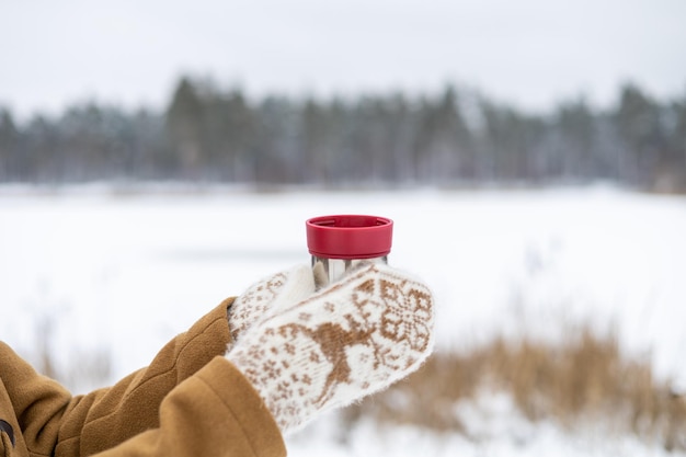 A girl in woolen mittens holds a red thermos with tea in her hands in a winter forest