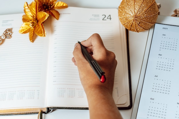 The girl writes in the diary next to the calendar for 2021 on tablet pc and wireless headphone, the concept of plans, wish list and to do list before Christmas