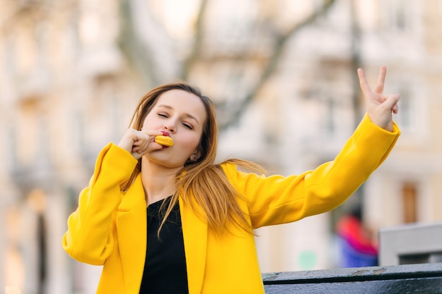 Girl in yellow coat eats yellow macaroon and shows a gesture