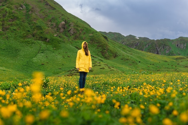 Girl in a yellow jacket stands in the middle of blooming alpine meadows in the green mountains