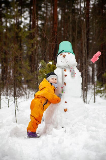 Girl in yellow jumpsuit in winter in forest near snowman