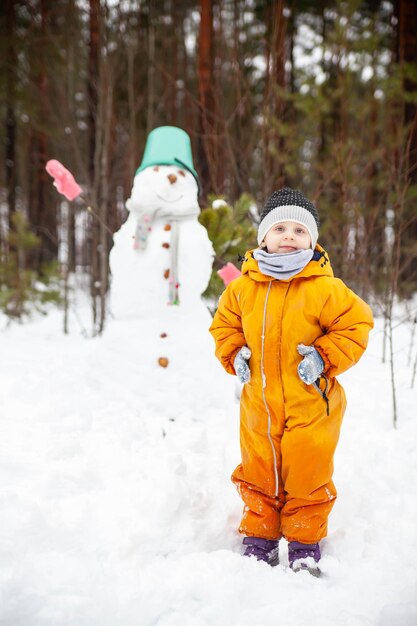 Girl in yellow jumpsuit in winter in forest near snowman