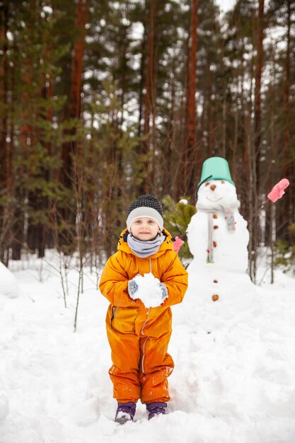 Girl in yellow jumpsuit in winter in forest near snowman