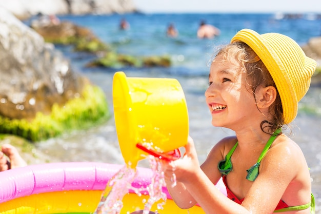 girl in yellow straw hat plays in outdoor near sea,  in an inflatable pool on the beach. 