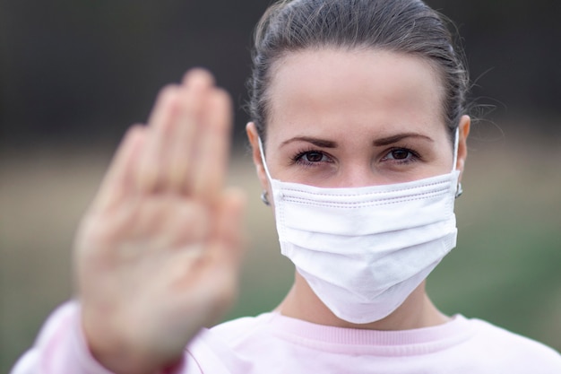 Girl, young woman in protective sterile medical mask on her face  outdoors, show palm, hand, stop no sign. Air pollution, virus, pandemic coronavirus concept. Covid-19