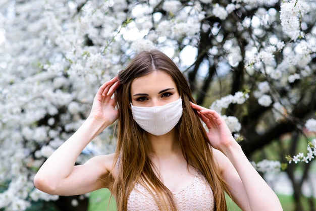 Girl, young woman in a protective sterile medical mask on her face in the spring garden. Air pollution, virus, pandemic coronavirus concept.