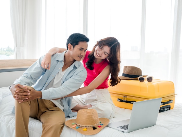 The girlfriend is begging and persuading her boyfriend to take a summer trip Young cute Asian woman and handsome man looking at the laptop display on bed with suitcase in bedroom Happy holiday