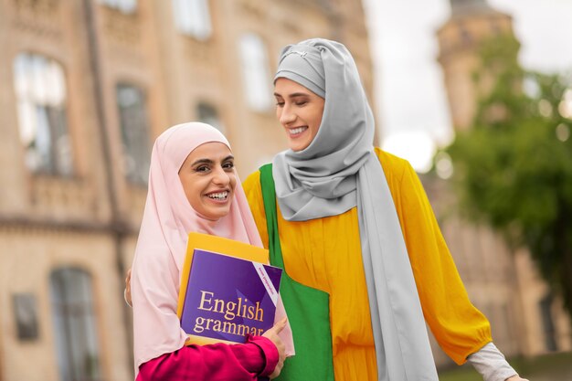 Girlfriend with friend. Beaming student wearing pink hijab holding English book standing near friend