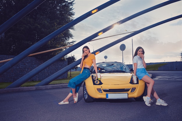 Girls in casual outfit eating hamburgers and holding beverages in paper cups while leaning on the hood of yellow car