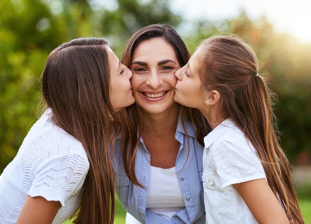 The girls of the family Portrait of a cheerful young woman receiving a kiss on each cheek from her young daughters outside in a park during the day