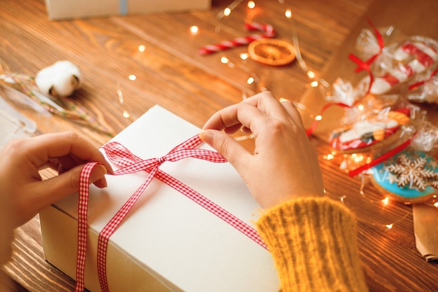 Girls hands in knitted sweater tie red ribbon bow on white gift box garland lights gingerbread and d...