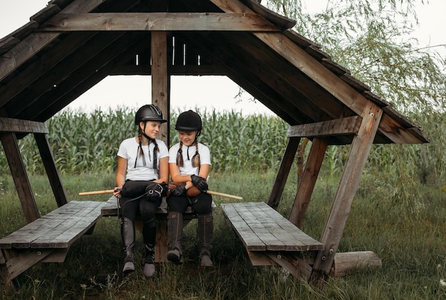 Girls jockeys sit on a wooden table under a canopy hold toys in their hands communicate Girlfriends are resting after a hobbyhorsing workout