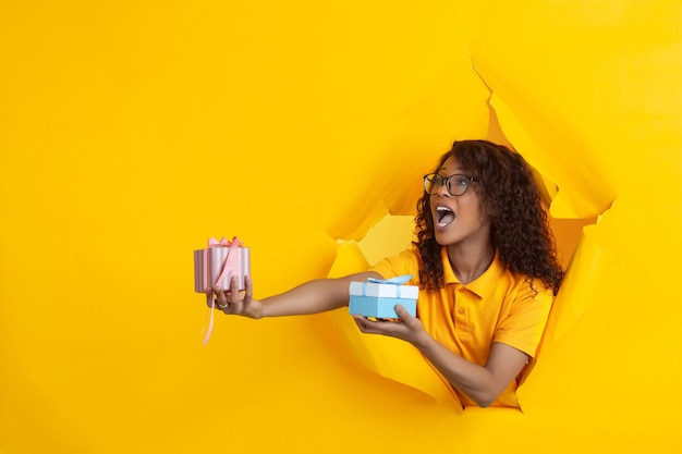 Giving gifts. Cheerful african-american young woman in torn yellow paper background, emotional, expressive. Breaking on, breakthrought. Concept of human emotions, facial expression, sales, ad.