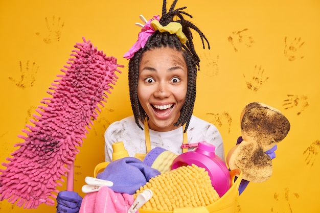 Glad Afro American woman with braids holds dirty sponge and mop looks happily at camera removes dirt everywhere isolated over yellow wall