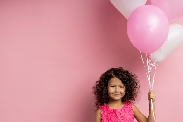 Glad birthday little girl with kinky dark hair holding flying balloons, smiling happily, wearing festive dress, standing with copy space for your text. Holiday, party concept.