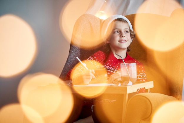 Glad boy in Santa hat looking into distance with dreamy face while writing letter to Santa near hot cocoa in room with bokeh during Christmas preparation