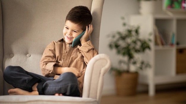 Glad caucasian little kid in casual calls by phone talk sits in armchair at spare time in living room interior