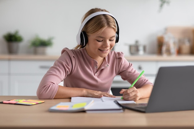 Glad european youngsters girl blonde pupil in headphones makes notes study at home at table