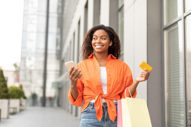 Glad happy millennial black lady in casual with smartphone credit card and a lot of packages