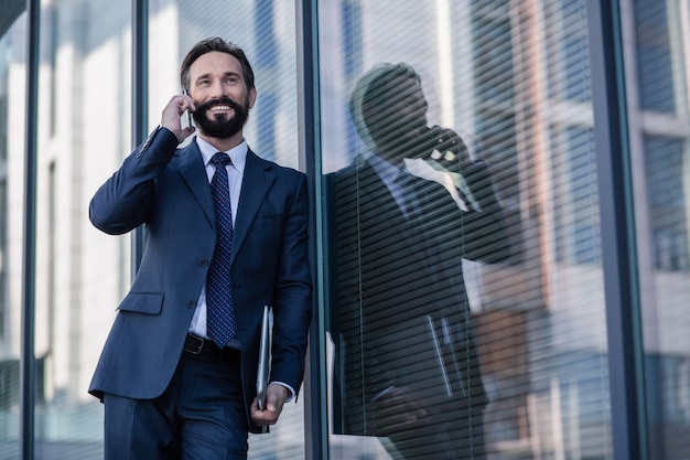 Glad to hear you. Cheerful smiling businessman having a pleasant conversation while standing against glass background