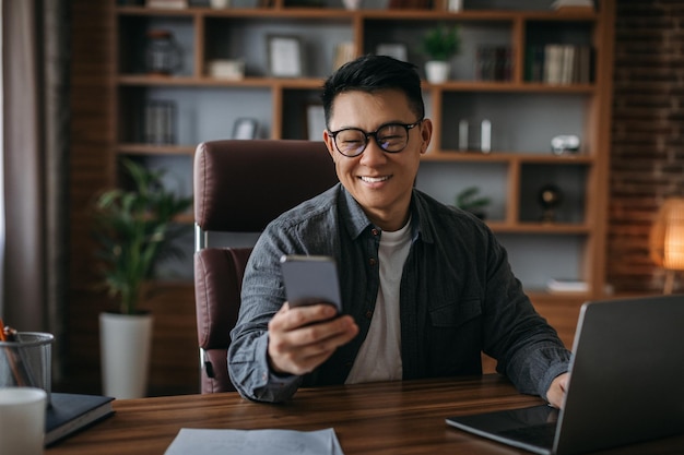 Glad mature korean male in glasses reads message on smartphone work on laptop in home office interior