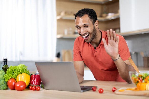 Glad millennial african american man at table with organic vegetables uses laptop waving hand for video call