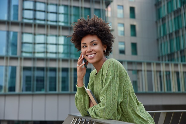 Glad positive woman with curly hair smiles happily has pleasant conversation makes call via smartphone holds notebook wears casual green jumper poses against urban setting Street lifestyle