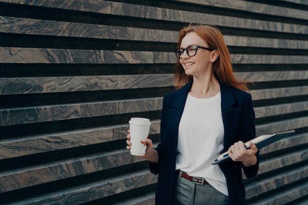 Glad redhead young woman enjoys life has successful day good mood drinks takeaway coffee holds digital tablet and notepad looks happily into distance wears formal clothes poses outdoor near wall