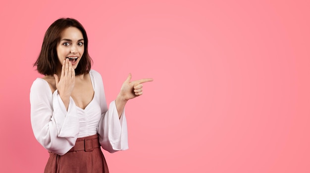 Glad shocked millennial caucasian brunette woman with open mouth pointing finger at free space