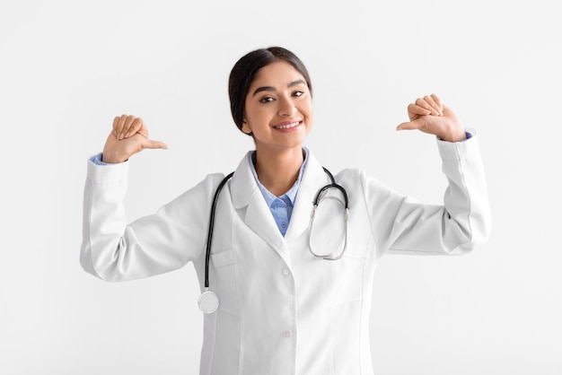 Glad smiling young indian female doctor in coat points fingers at herself isolated on white background studio