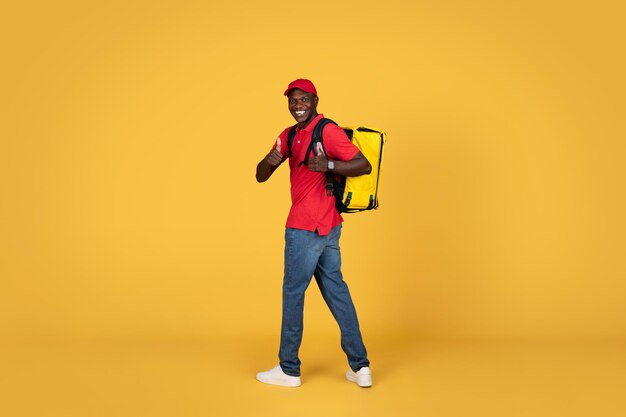 Photo glad young african american delivery man in uniform and cap with big backpack show thumbs up sign