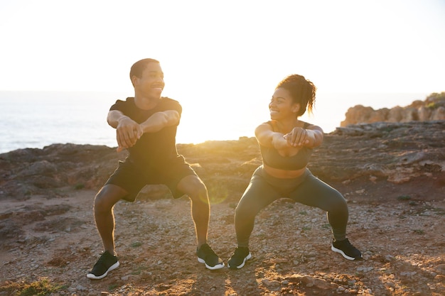 Glad young african american girlfriend and man in sportswear squatting on ocean beach enjoy morning